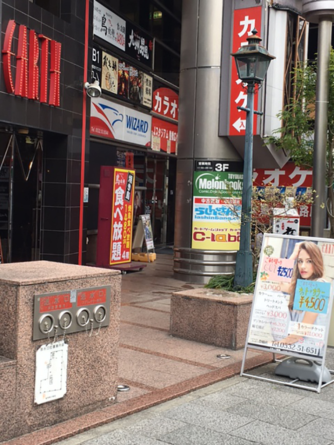 横断歩道を渡りつつ進むと柱に看板があります。（カラオケ側の自動ドアからビルの中へ）